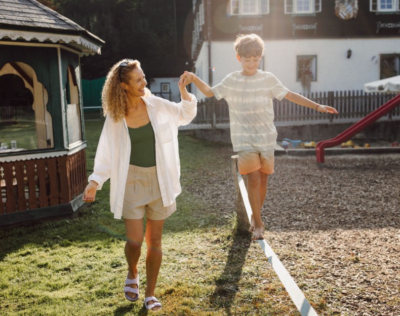 Ein Junge balanciert auf einem Brett im Freien, während eine Frau seine Hand hält