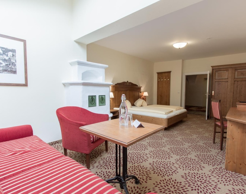 Helles Zimmer mit Bett, rotem Sessel, Tisch und traditionellem Ofen an der Wand im Ennshof, Hotel Resl in Flachau