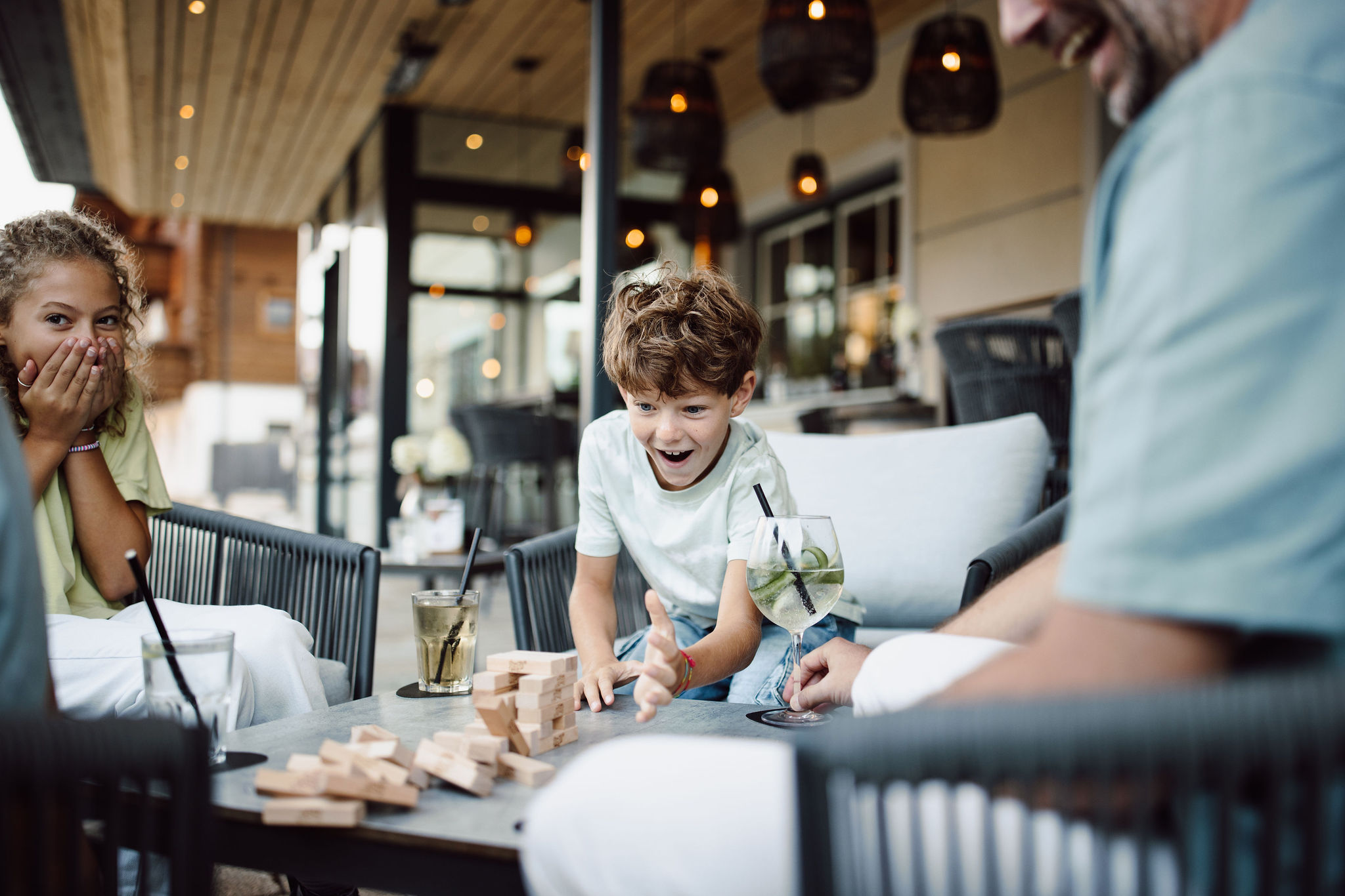 Ein lachender Junge spielt Jenga draußen an einem Tisch mit einem Getränk daneben