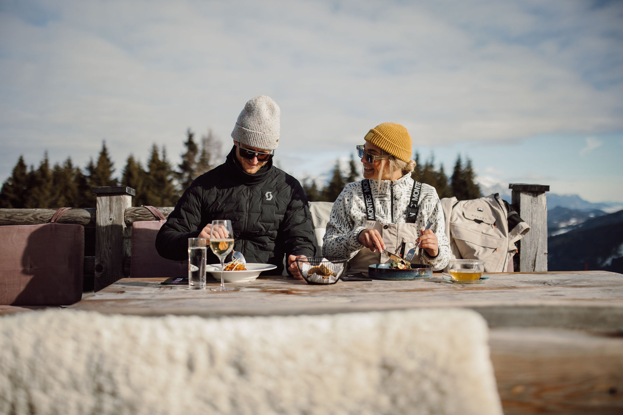 Zwei Personen im Wintermantel essen draußen an einem Holztisch mit Getränken und Snacks