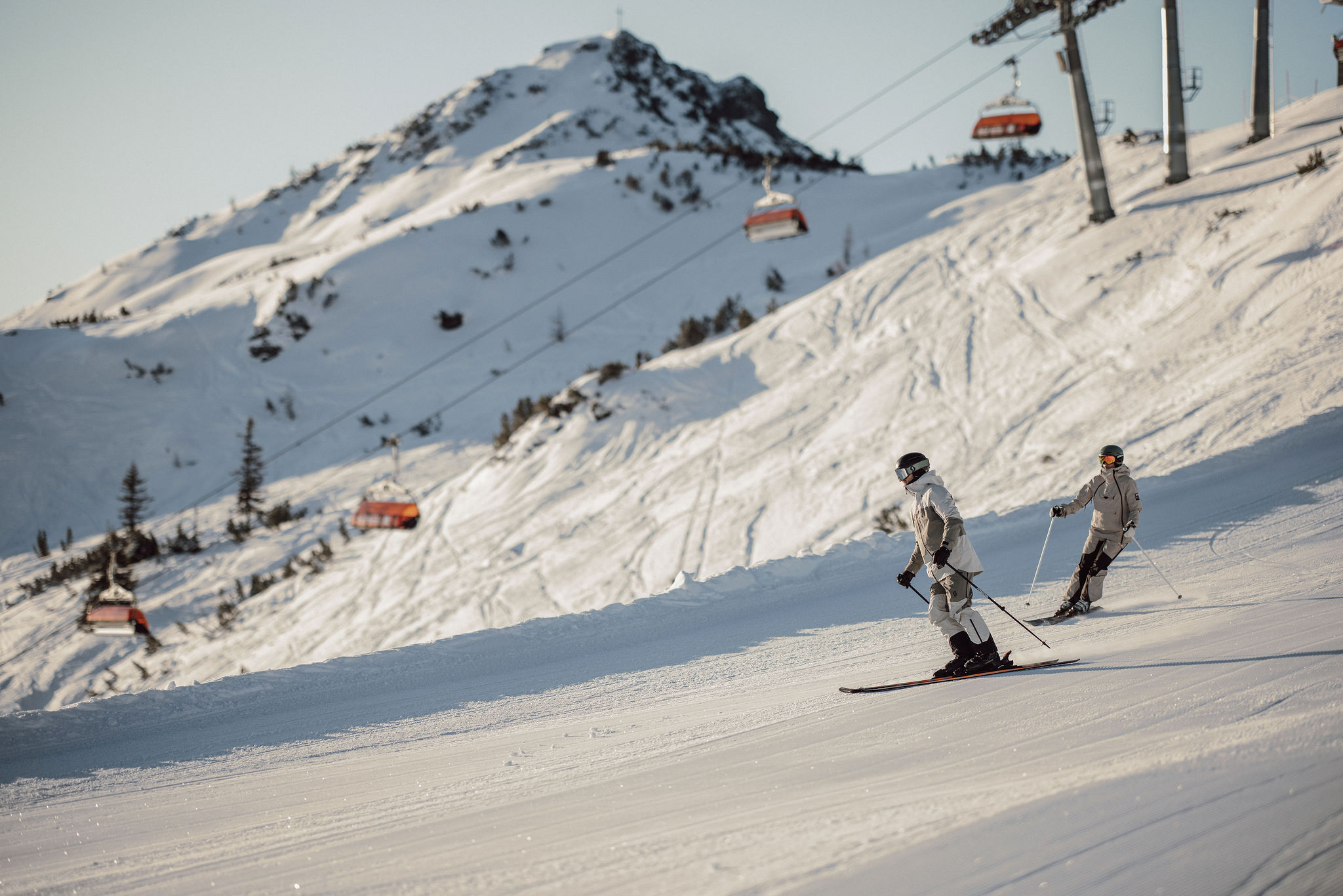 Zwei Skifahrer auf einer schneebedeckten Piste mit roten Gondeln im Hintergrund