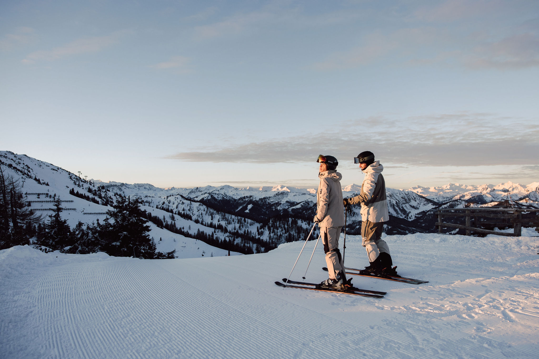 Two skiers stand on a snowy hill overlooking distant mountains at sunset