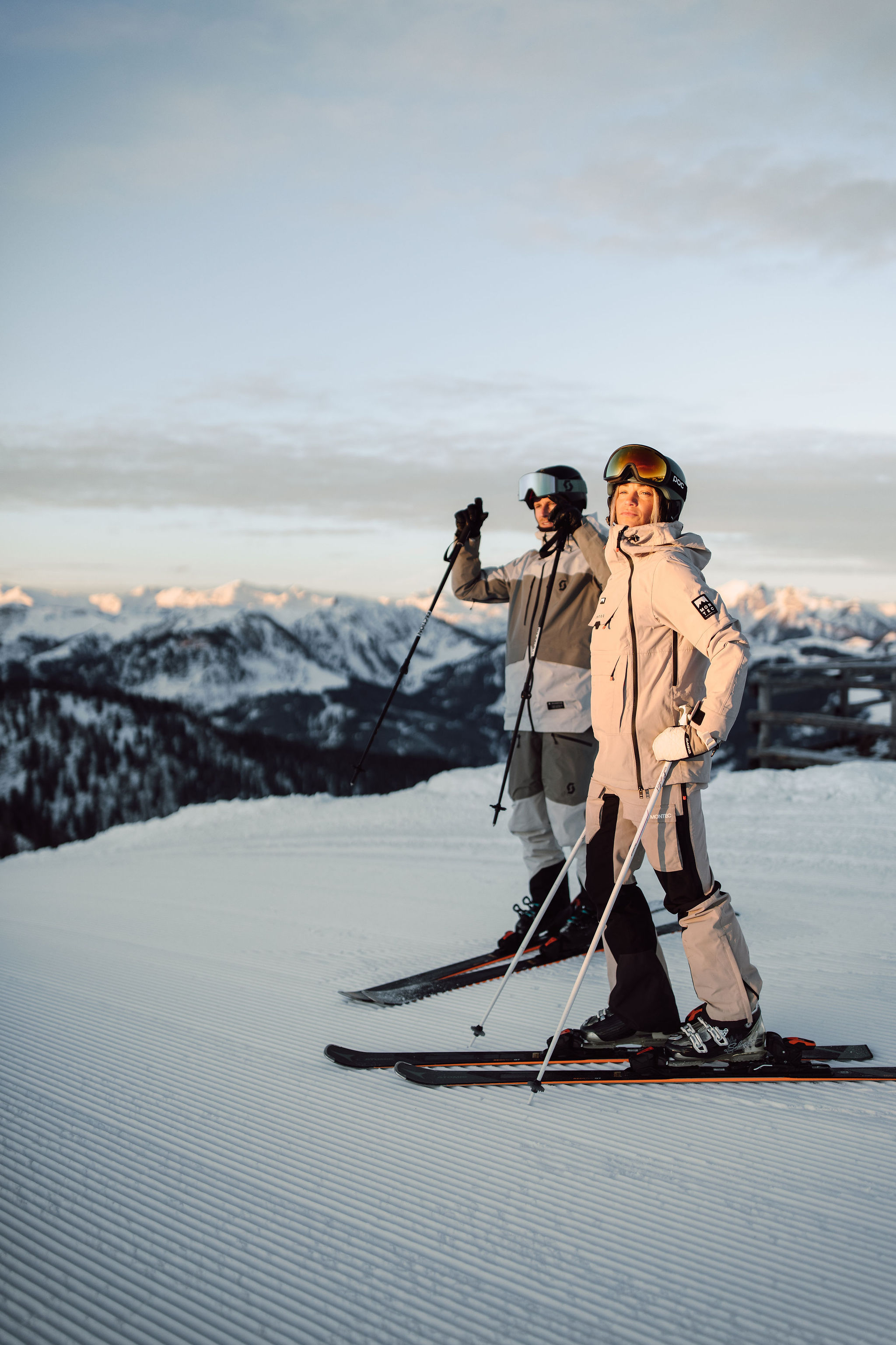 Two skiers stand on a snowy mountain with a scenic view in the background
