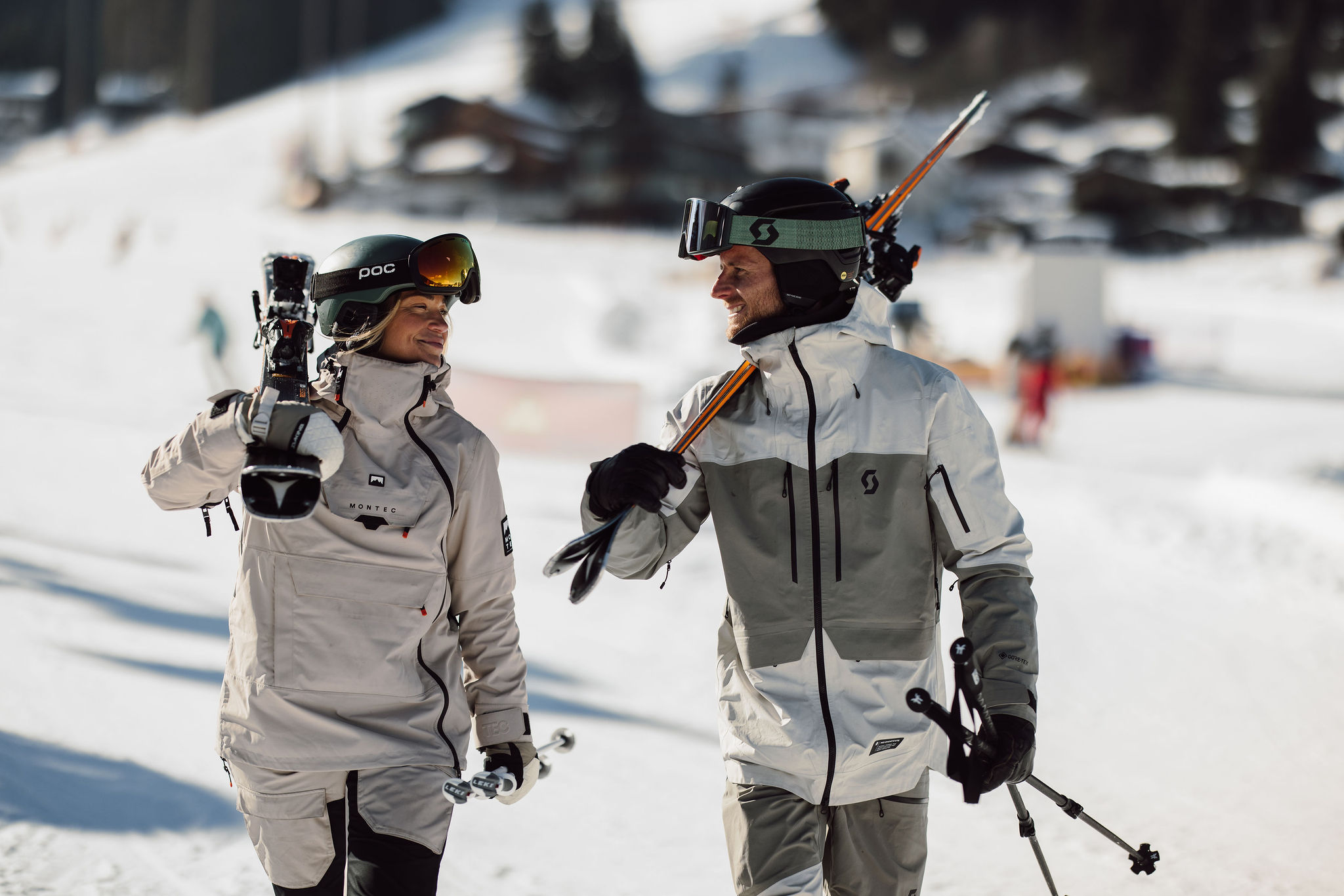 Two people in winter gear chat while carrying skis on a snowy slope