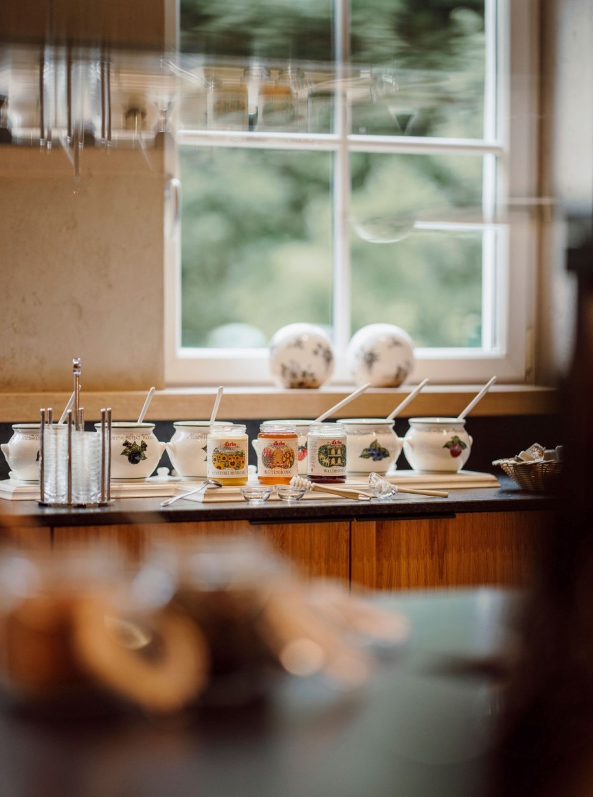 Mehrere Marmeladengläser und weiße Keramiktöpfe mit Löffeln auf einer Theke vor einem Fenster