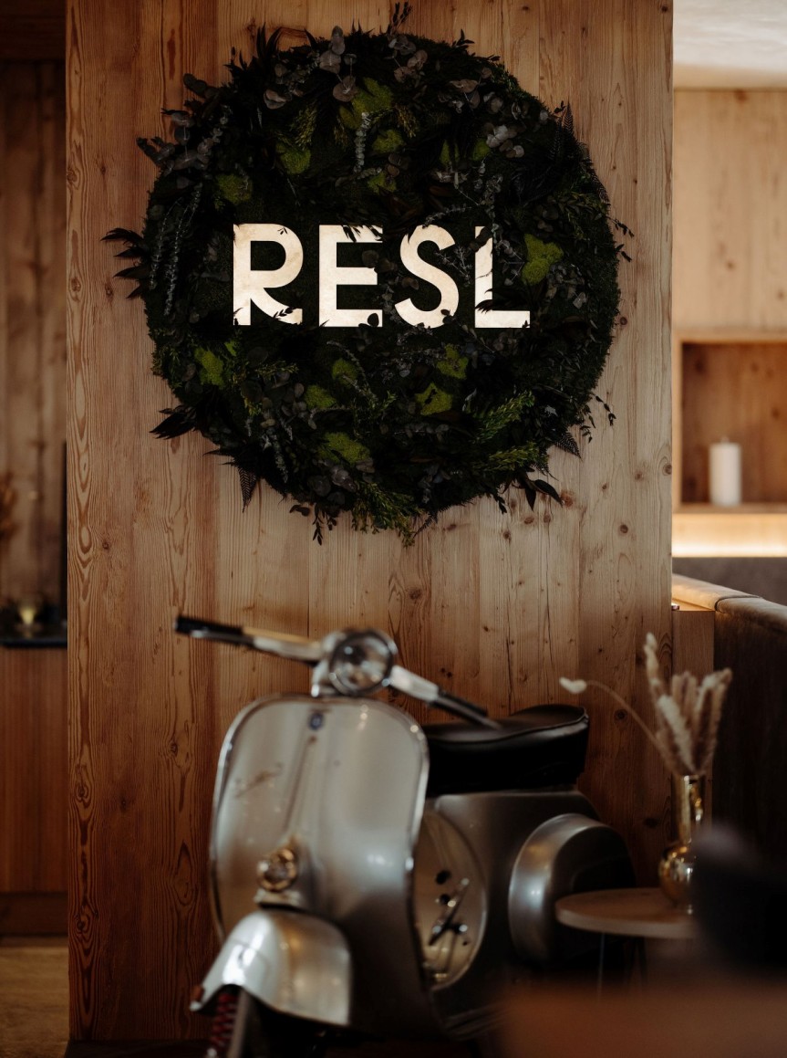 Ein alter Roller steht vor einer Holzwand mit Pflanzenkranz und dem Wort "RESL"