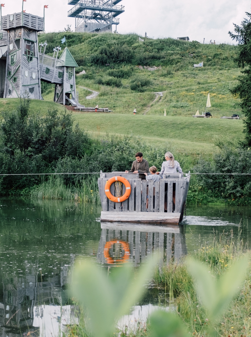 Kinder und Familien fahren auf einem Holzflo&szlig; &uuml;ber einen kleinen See in einer gr&uuml;nen Landschaft