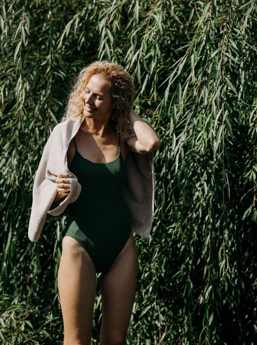 Frau im Badeanzug vor grüner Pflanzenwand, lächelt mit Hand im Haar