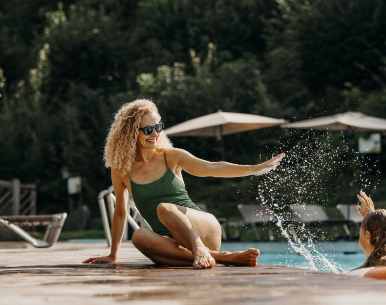 Eine Frau im Badeanzug sitzt lachend am Poolrand und spielt mit ihren Kindern.