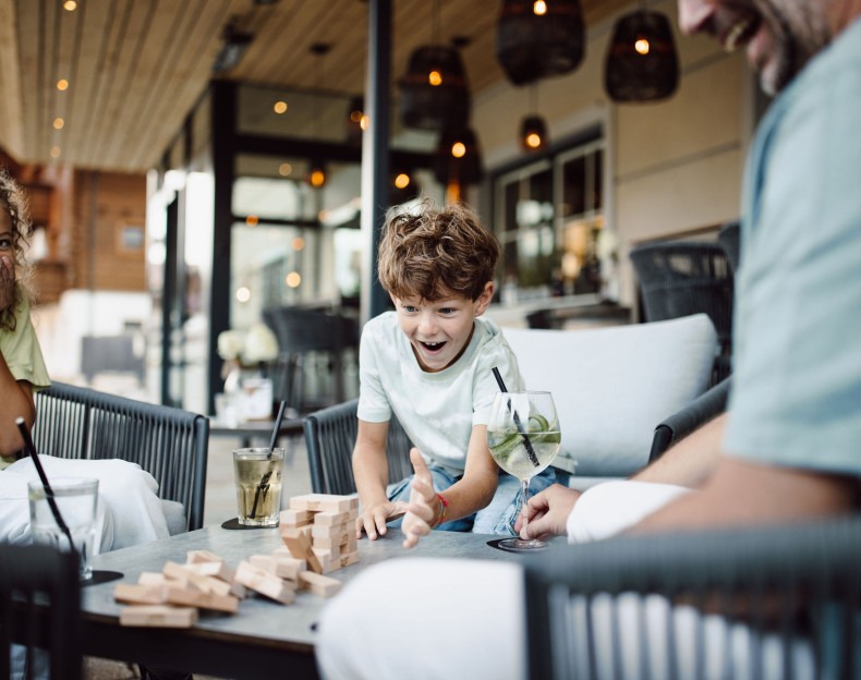 Ein Junge spielt begeistert Jenga mit seiner Familie auf einer Terrasse, auf der Getränke bereitstehen.