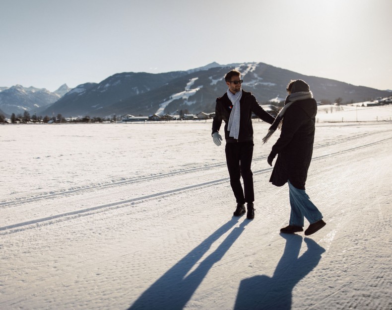 Zwei Personen spazieren auf schneebedecktem Feld vor Bergkulisse im Sonnenlicht