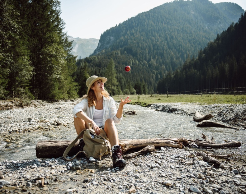 Frau sitzt am Bach in den Bergen und wirft einen Apfel in die Luft
