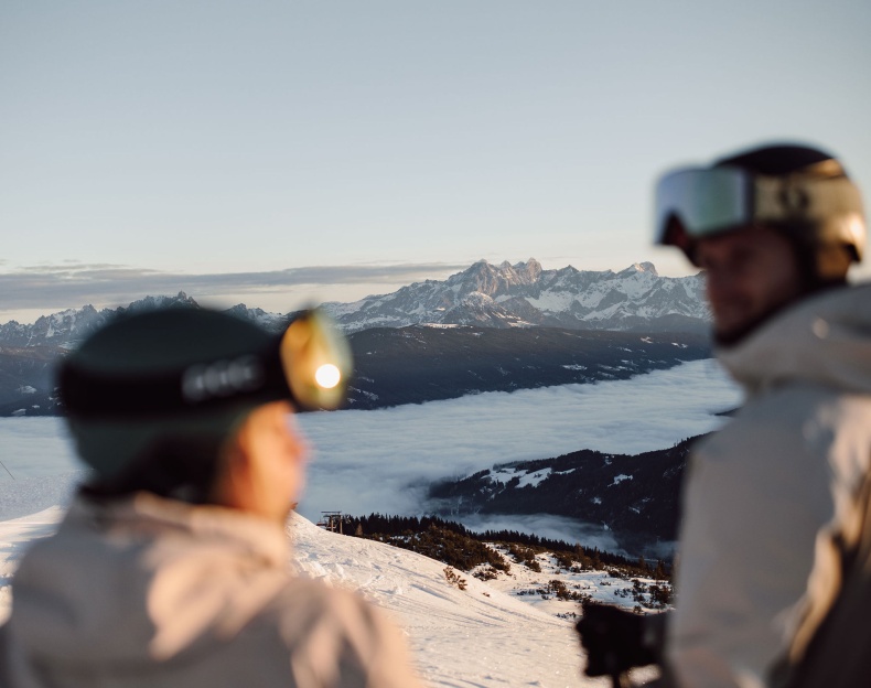 Paar spaziert bei sonnigem Wetter durch eine verschneite Landschaft mit Bergen im Hintergrund