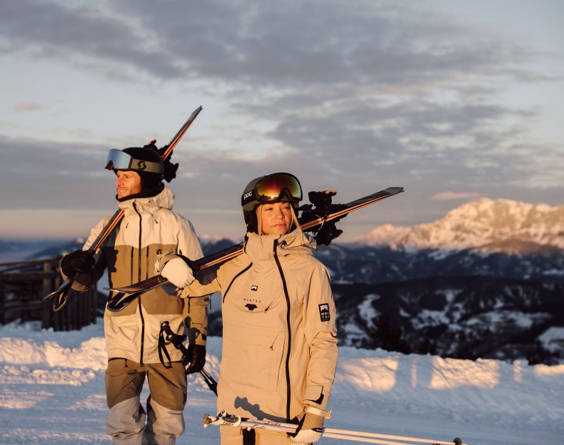 Zwei Personen stehen auf der Piste am Berg und die Sonne scheint ihnen ins Gesicht
