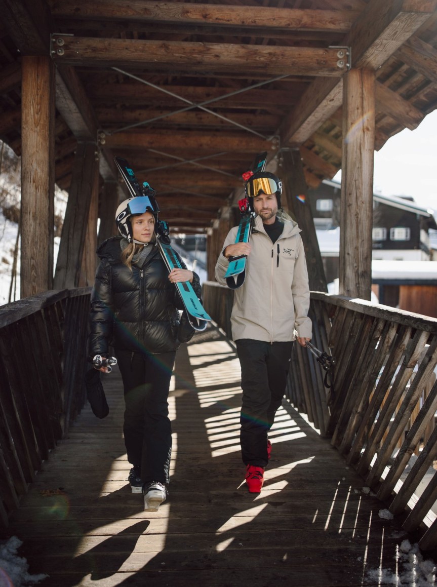 Zwei Personen tragen Skier auf einer überdachten Holzbrücke