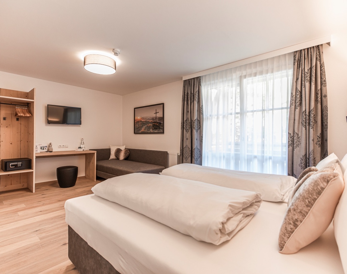 Helles Hotelzimmer mit Doppelbett, Sofa, Fernseher und großem Fenster mit Vorhängen